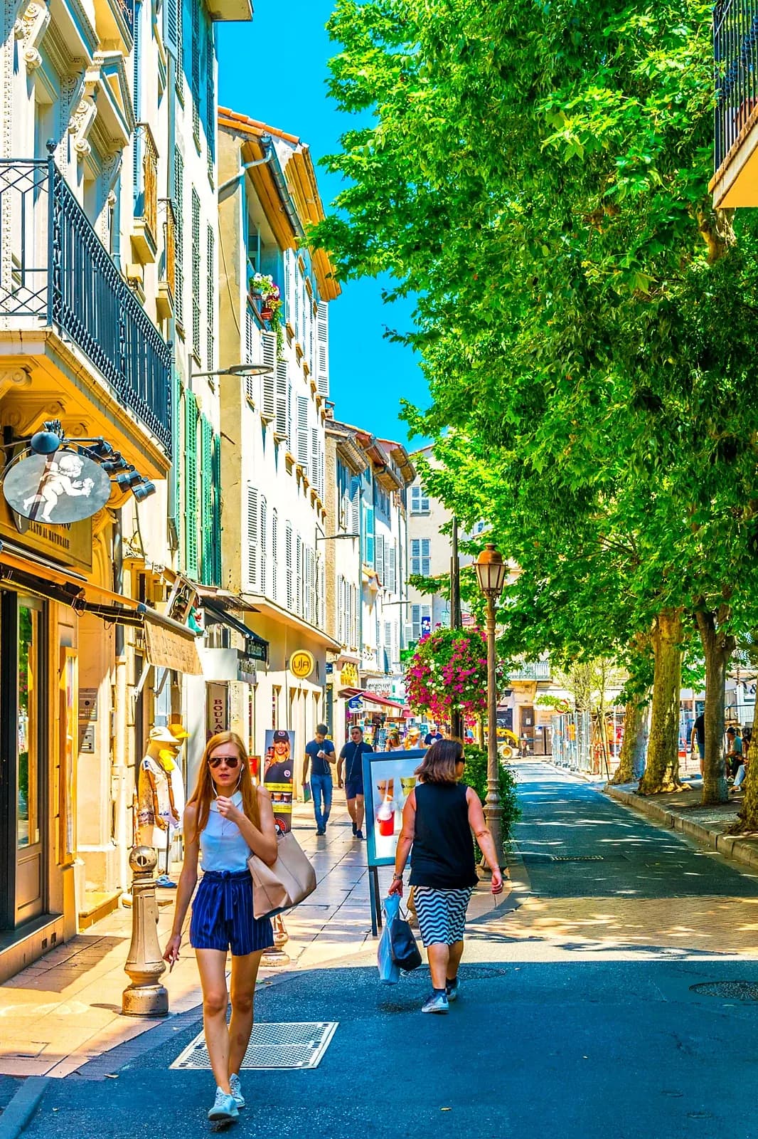 Shoppers on a plane-tree-lined shopping street in Antibes, shopfronts and flowers